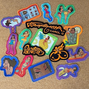May include: A collection of embroidered patches with various designs. The patches feature illustrations of women in different poses, tropical themes, and text including "Tropi G", "Karol G", and "Parasitic". The patches have colorful borders and detailed artwork.