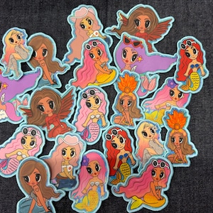 May include: A collection of colorful mermaid patches with various hairstyles, outfits, and accessories. Each patch features a cartoon mermaid with a unique design, including sunglasses, wings, and different colored hair. The patches are outlined in light blue.