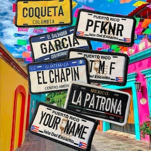 May include: A collection of embroidered license plate patches with various names and country affiliations. The patches feature text like "COQUETA COLOMBIA", "GARCIA CENTROAME", and "YOUR NAME". The background includes colorful buildings and festive decorations.