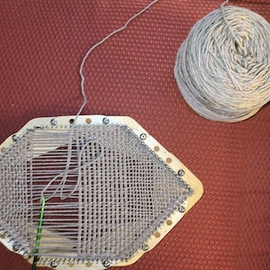 May include: A wooden loom with a gray yarn being woven into a pattern. A ball of gray yarn sits on a red surface.