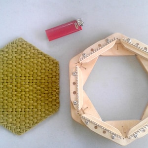 May include: A wooden hexagonal loom with six sides and a woven green fabric mat. The loom has a series of pegs on each side and is used for creating woven fabric.