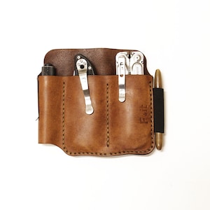 Könnte beinhalten: Ein braunes Lederetui für Multitools mit drei Fächern. Das Etui hat einen genähten Rand und den Text "F.A.L." auf der Vorderseite geprägt. Das Etui enthält ein Multitool, ein Taschenmesser und einen Stift.