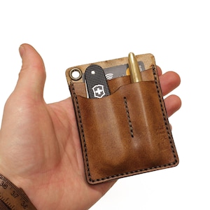 Könnte beinhalten: Ein brauner Leder-Taschenorganizer mit zwei Steckplätzen zum Tragen eines Stifts und eines kleinen Taschenmessers. Der Organizer hat einen genähten Rand und eine kleine Metallschlaufe.