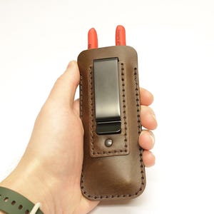 Op de afbeelding: Bruin leren radioholster met een zwarte metalen riemclip. Het holster heeft een gestikt ontwerp en een bruine afwerking.