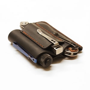 Peut inclure: Un porte-outils multifonction en cuir noir avec des détails de couture marron. Le porte-outils est rempli d'une variété d'outils, y compris une lampe de poche, un multi-outil et un couteau.