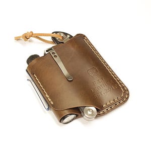 May include: A brown leather pocket organizer with a stitched edge and a metal clip. The organizer has a small flashlight and a multi-tool inside. The leather is embossed with the text "F.J.T. Leather Est. 2017."