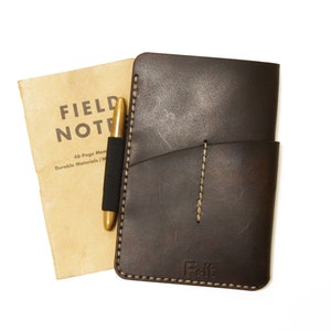 Può includere: Una copertina per quaderno in pelle marrone con una penna e un piccolo quaderno che dice "FIELD NOTES 48-Page Memo Durable Materials / Made in USA" all'interno. La copertina ha una tasca sul davanti e un bordo cucito.