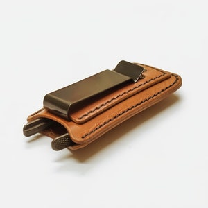 May include: A brown leather wallet with a metal clip and a stitched design. The wallet is closed and lying on a white surface.