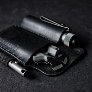 May include: A black leather carrying case with three compartments for tools. The case has a stitched edge and the word "Leathercraft" embossed on the front. The case is holding a small silver tool, a black and silver multi-tool, and a black flashlight.