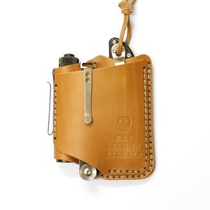 May include: A tan leather knife sheath with a metal clip and a loop for a lanyard. The sheath has a stitched edge and the text "FIT LEATHER EST. 2017" is embossed on the front.