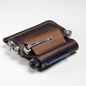 May include: A brown leather multi-tool pouch with a brown leather flap and a silver multi-tool inside. The pouch has a blue metal pen holder on the side.