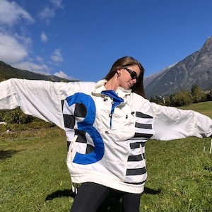 Könnte beinhalten: Vintage Anorak-Jacke in Weiß und Blau mit einer großen blauen Zahl 3 auf der Vorderseite, akzentuiert mit schwarz-weiß karierten und schwarz gestreiften Details. Die Jacke hat einen hohen Kragen und einen Kordelzug am Saum. Sportlicher Retro-Stil.