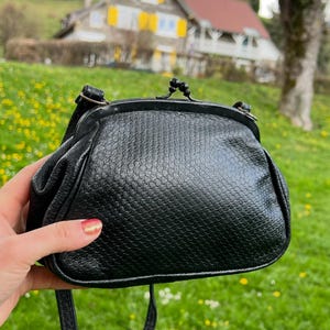 K&ouml;nnte beinhalten: Schwarze, strukturierte Umh&auml;ngetasche mit Kussverschluss. Die Tasche hat einen langen, d&uuml;nnen Riemen und eine runde Form. Die Au&szlig;enseite hat ein schuppenartiges Muster. Die Tasche wird vor einem Hintergrund aus gr&uuml;nem Gras und einem Haus gehalten.