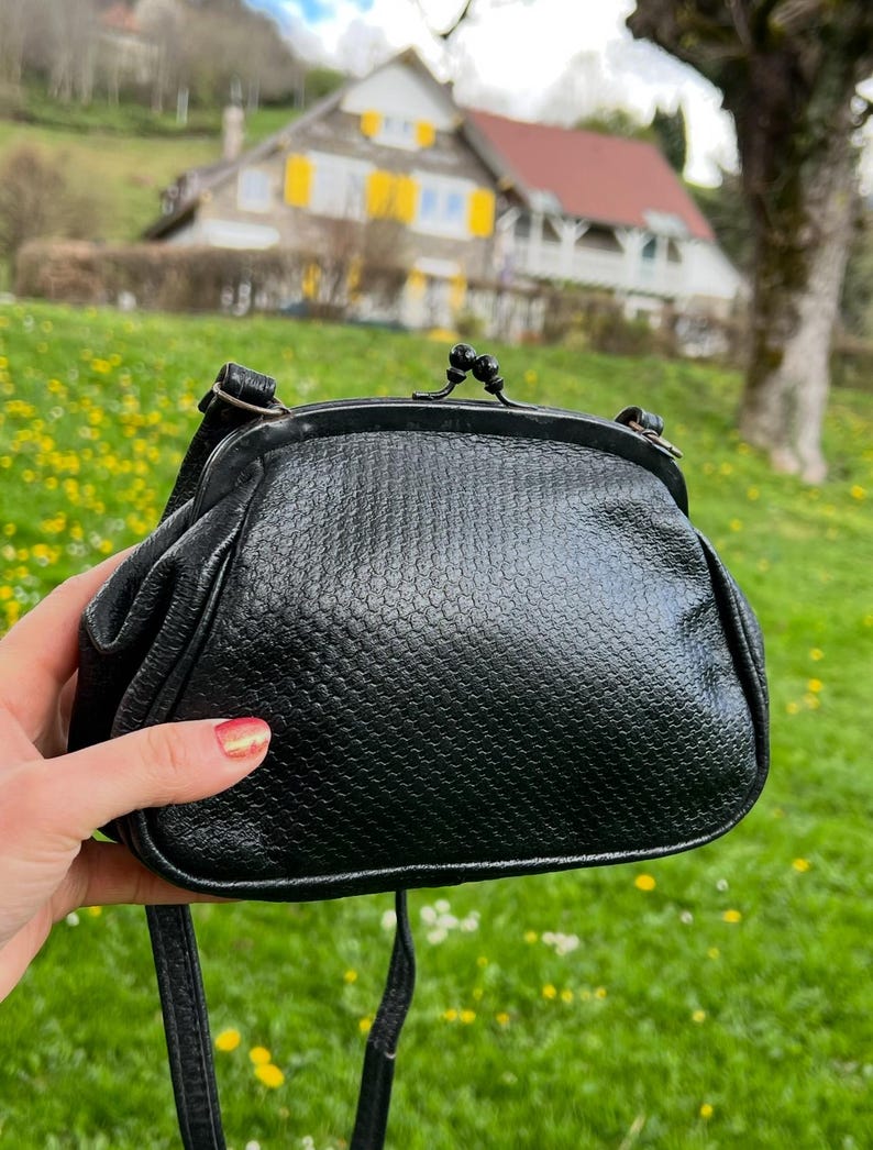 K&ouml;nnte beinhalten: Schwarze, strukturierte Umh&auml;ngetasche mit Kussverschluss. Die Tasche hat einen langen, d&uuml;nnen Riemen und eine runde Form. Die Au&szlig;enseite hat ein schuppenartiges Muster. Die Tasche wird vor einem Hintergrund aus gr&uuml;nem Gras und einem Haus gehalten.