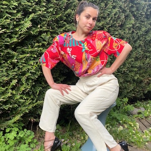 May include: A red, short-sleeved top with a colorful floral pattern, paired with cream-colored pants. The outfit is completed with black high-heeled sandals. The person is sitting on a blue stool in a garden setting.