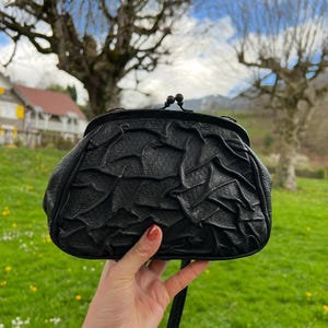 K&ouml;nnte beinhalten: Schwarze Lederhandtasche mit strukturiertem, ger&uuml;schtem Design. Die Tasche hat einen Schnallenverschluss und einen langen Riemen. Der Hintergrund zeigt eine gr&uuml;ne Wiese, B&auml;ume und einen blauen Himmel mit Wolken.