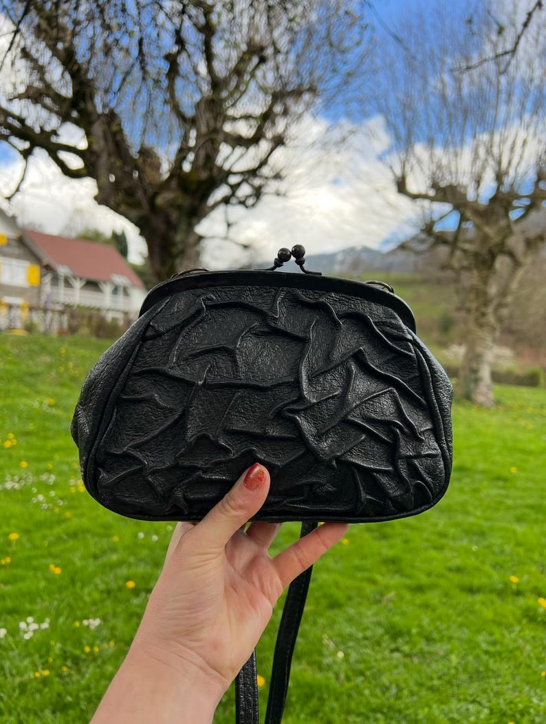 K&ouml;nnte beinhalten: Schwarze Lederhandtasche mit strukturiertem, ger&uuml;schtem Design. Die Tasche hat einen Schnallenverschluss und einen langen Riemen. Der Hintergrund zeigt eine gr&uuml;ne Wiese, B&auml;ume und einen blauen Himmel mit Wolken.