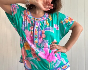 May include: A green, short-sleeved blouse with a floral print featuring pink, purple, and orange flowers. The blouse has a button-up front and a white trim around the neckline.
