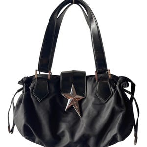 May include: A black nylon handbag featuring two structured shoulder straps, a central flap closure adorned with a prominent silver-toned metallic star emblem, and drawstring side details with metallic hardware accents on a soft, gathered body.