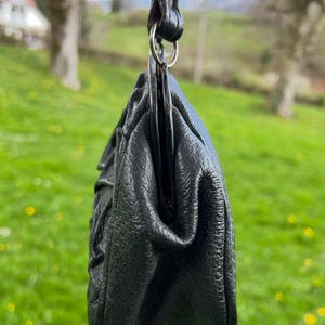 K&ouml;nnte beinhalten: Eine schwarze Lederhandtasche mit strukturierter Oberfl&auml;che, aufgeh&auml;ngt an einem passenden Riemen und einem silberfarbenen Ring. Das Design der Tasche weist gerafften Stoff auf, der eine weiche, abgerundete Form erzeugt. Der Hintergrund ist ein verschwommenes gr&uuml;nes Feld.
