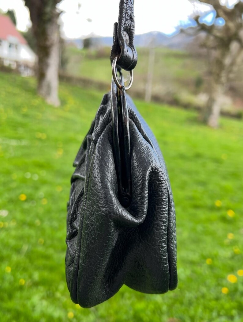 K&ouml;nnte beinhalten: Eine schwarze Lederhandtasche mit strukturierter Oberfl&auml;che, aufgeh&auml;ngt an einem passenden Riemen und einem silberfarbenen Ring. Das Design der Tasche weist gerafften Stoff auf, der eine weiche, abgerundete Form erzeugt. Der Hintergrund ist ein verschwommenes gr&uuml;nes Feld.