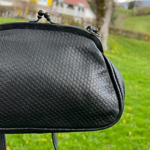 K&ouml;nnte beinhalten: Schwarze Vintage-Handtasche mit strukturierter Oberfl&auml;che. Die Tasche hat einen Schnappverschluss und einen langen Riemen. Der Hintergrund zeigt eine verschwommene Szene im Freien mit Gr&uuml;n und Geb&auml;uden.