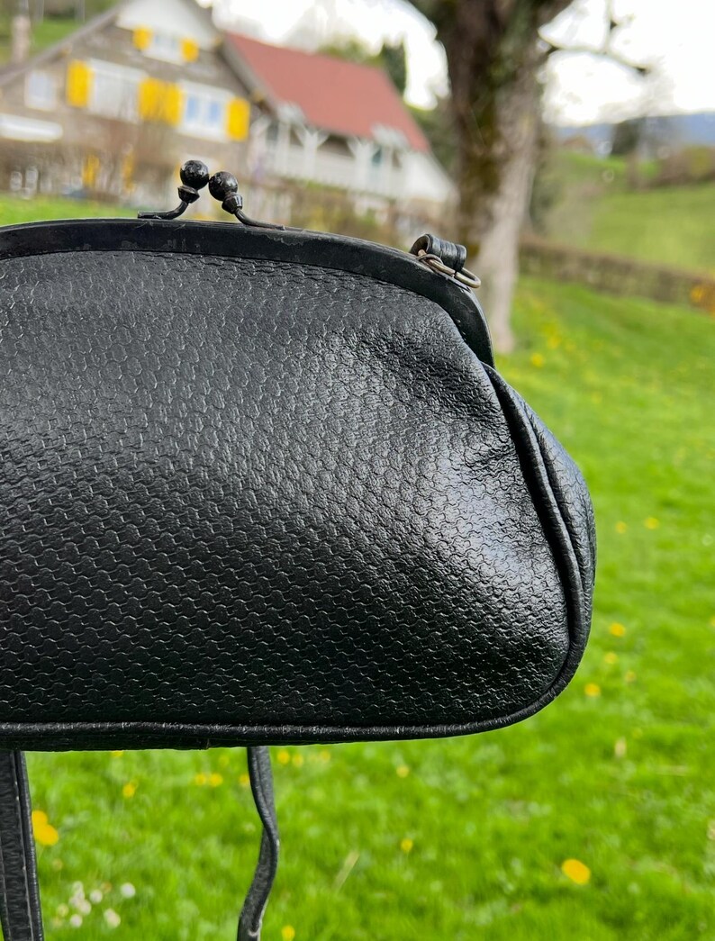 K&ouml;nnte beinhalten: Schwarze Vintage-Handtasche mit strukturierter Oberfl&auml;che. Die Tasche hat einen Schnappverschluss und einen langen Riemen. Der Hintergrund zeigt eine verschwommene Szene im Freien mit Gr&uuml;n und Geb&auml;uden.