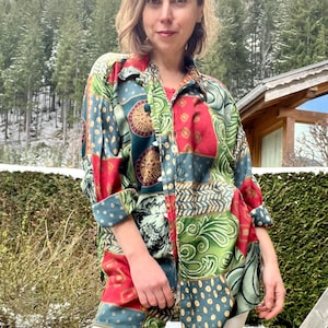Peut inclure: Une chemise patchwork avec des panneaux aux motifs rouges, verts et bleu canard. La chemise a des manches longues et un col. Le motif comprend des motifs floraux et géométriques. La personne porte un pantalon blanc.