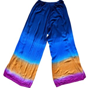 May include: Wide-leg pants with a vibrant, tie-dye effect. The color gradient transitions from deep blue at the top to shades of orange and purple at the hem. The pants are made of a soft, flowing fabric.