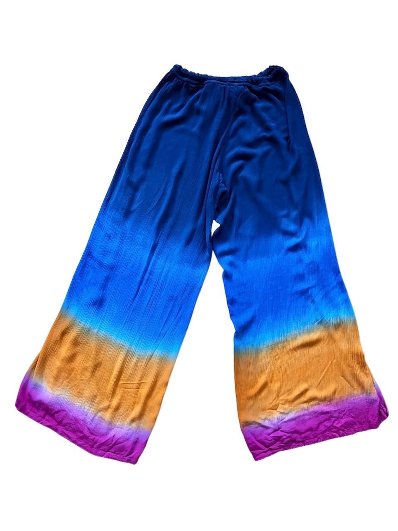 May include: Wide-leg pants with a vibrant, tie-dye effect. The color gradient transitions from deep blue at the top to shades of orange and purple at the hem. The pants are made of a soft, flowing fabric.