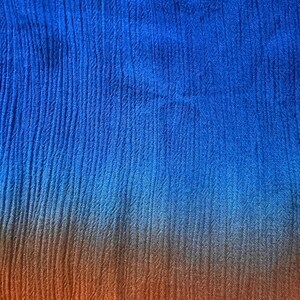 May include: Close-up of a textured fabric with a color gradient. The top portion is a vibrant blue, transitioning to a warm orange at the bottom. The fabric has vertical, subtle ribbed lines.