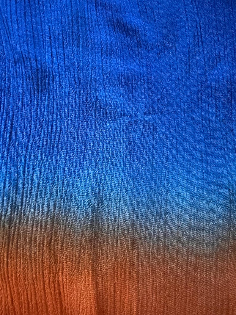 May include: Close-up of a textured fabric with a color gradient. The top portion is a vibrant blue, transitioning to a warm orange at the bottom. The fabric has vertical, subtle ribbed lines.