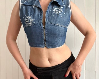 May include: A blue denim vest with a zipper closure and white stitching. The vest has a cropped length and is distressed with holes and frayed edges.