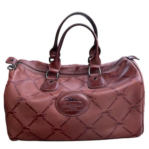 May include: A brown leather duffel bag with two top handles and a zip closure. The bag features a repeating pattern of a horse and rider, with a leather patch that reads "LONGCHAMP PARIS MADE IN FRANCE".