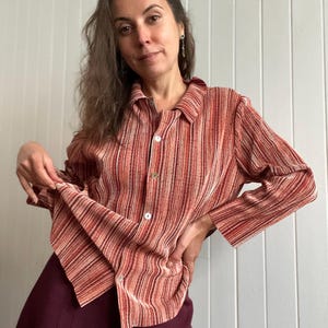 Pode incluir: Camisa listrada verticalmente em tons de vermelho, laranja e branco, com gola e botões. As mangas compridas são combinadas com uma saia bordô. A camisa é feita de um tecido leve.