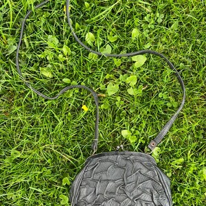 K&ouml;nnte beinhalten: Eine schwarze, strukturierte Vintage-Handtasche mit einem langen, d&uuml;nnen Riemen. Die Handtasche hat einen Schnappverschluss und befindet sich vor einem Hintergrund aus gr&uuml;nem Gras.