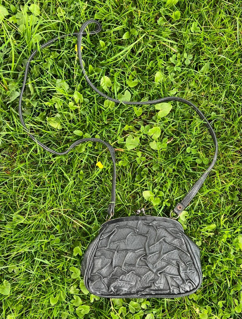 K&ouml;nnte beinhalten: Eine schwarze, strukturierte Vintage-Handtasche mit einem langen, d&uuml;nnen Riemen. Die Handtasche hat einen Schnappverschluss und befindet sich vor einem Hintergrund aus gr&uuml;nem Gras.