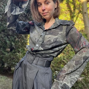 Puede incluir: Una camisa oscura estampada con mangas con volantes y un cinturón ancho. La camisa presenta un estampado con tonos grises claros y oscuros, y la falda es de diseño a rayas grises oscuras.