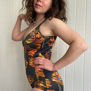 以下が含まれることがあります： オレンジ、イエロー、ブルーのアクセントがついた花柄のブラックのワンピース水着。水着は深いVネックと細いストラップです。