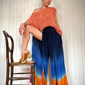 May include: An orange crochet top and navy blue, blue, orange, and pink ombre palazzo pants. The pants have a wide leg and a drawstring waist. The model is wearing brown clogs with a wooden sole. A wooden chair is in the background.