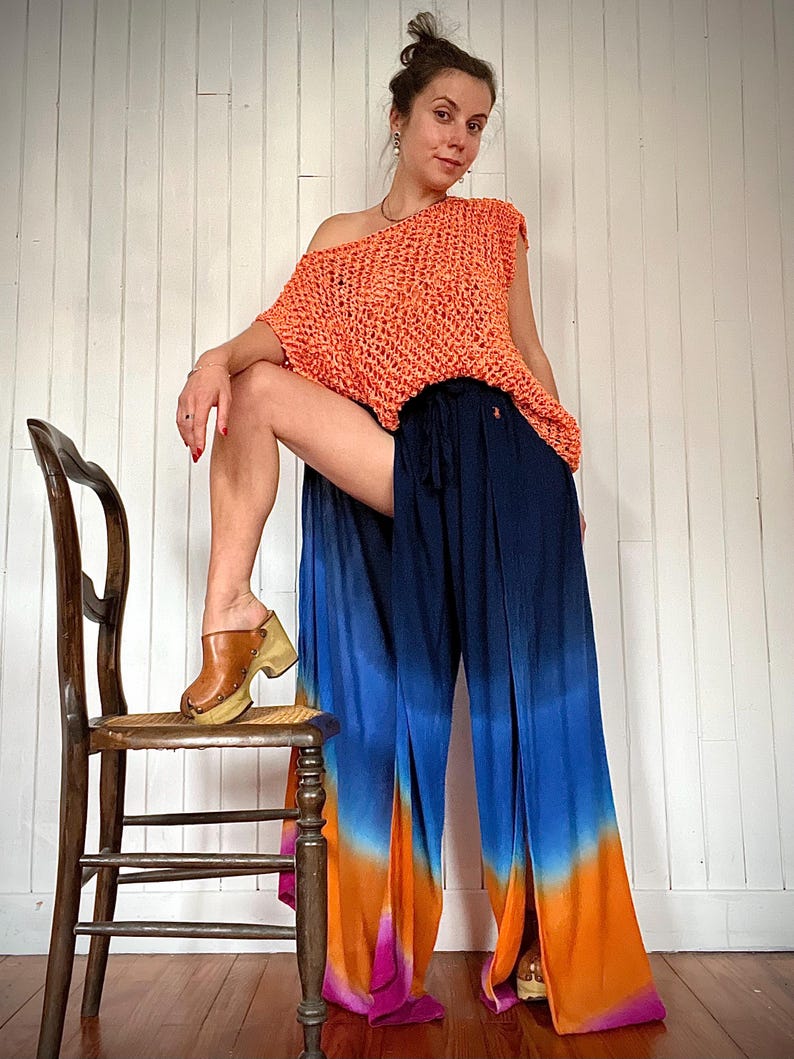 May include: An orange crochet top and navy blue, blue, orange, and pink ombre palazzo pants. The pants have a wide leg and a drawstring waist. The model is wearing brown clogs with a wooden sole. A wooden chair is in the background.