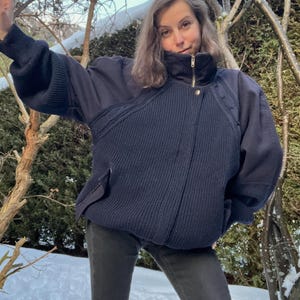 May include: A navy blue jacket with a high collar and gold zip. The jacket has a ribbed knit front panel and a relaxed fit. The sleeves are a darker navy and have a slight puff. The person is wearing dark grey trousers.