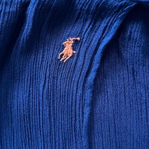 May include: Close-up of a textured, royal blue fabric with a small, embroidered light brown polo player logo. The fabric has vertical pleats and a subtle sheen, suggesting a garment.
