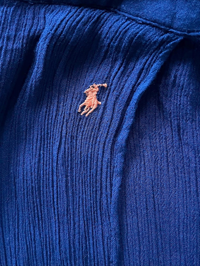 May include: Close-up of a textured, royal blue fabric with a small, embroidered light brown polo player logo. The fabric has vertical pleats and a subtle sheen, suggesting a garment.