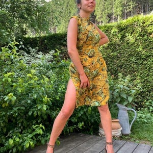 Può includere: Un abito giallo senza maniche con un motivo verde e marrone. L'abito ha uno scollo alto e uno spacco sulla coscia. La persona indossa tacchi leopardati. Lo sfondo comprende fogliame verde e un terrazzo in legno.