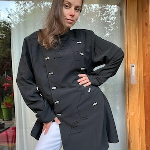 Puede incluir: Una chaqueta negra de manga larga con un diseño único. La chaqueta presenta una serie de pequeños acentos rectangulares y detalles de texto sutiles. La chaqueta se combina con pantalones blancos. El estilo general es moderno y elegante.