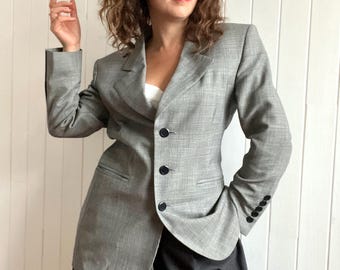 May include: A grey and white patterned blazer with a single button fastening and a black pocket on the left side. The blazer is worn over a white top and black trousers.