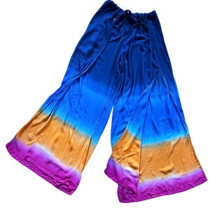 May include: Blue, orange, and purple tie-dye palazzo pants. The wide-leg pants have a drawstring waist and a split design. The fabric has a textured appearance and a gradient color scheme, transitioning from blue to orange and purple.