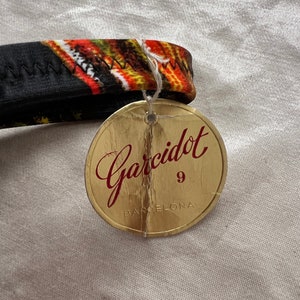 以下が含まれることがあります： 赤文字で「Garcidot 9 BARCELONA」と書かれた金色のタグ。タグはジグザグ模様の黒とオレンジの生地に付けられています。