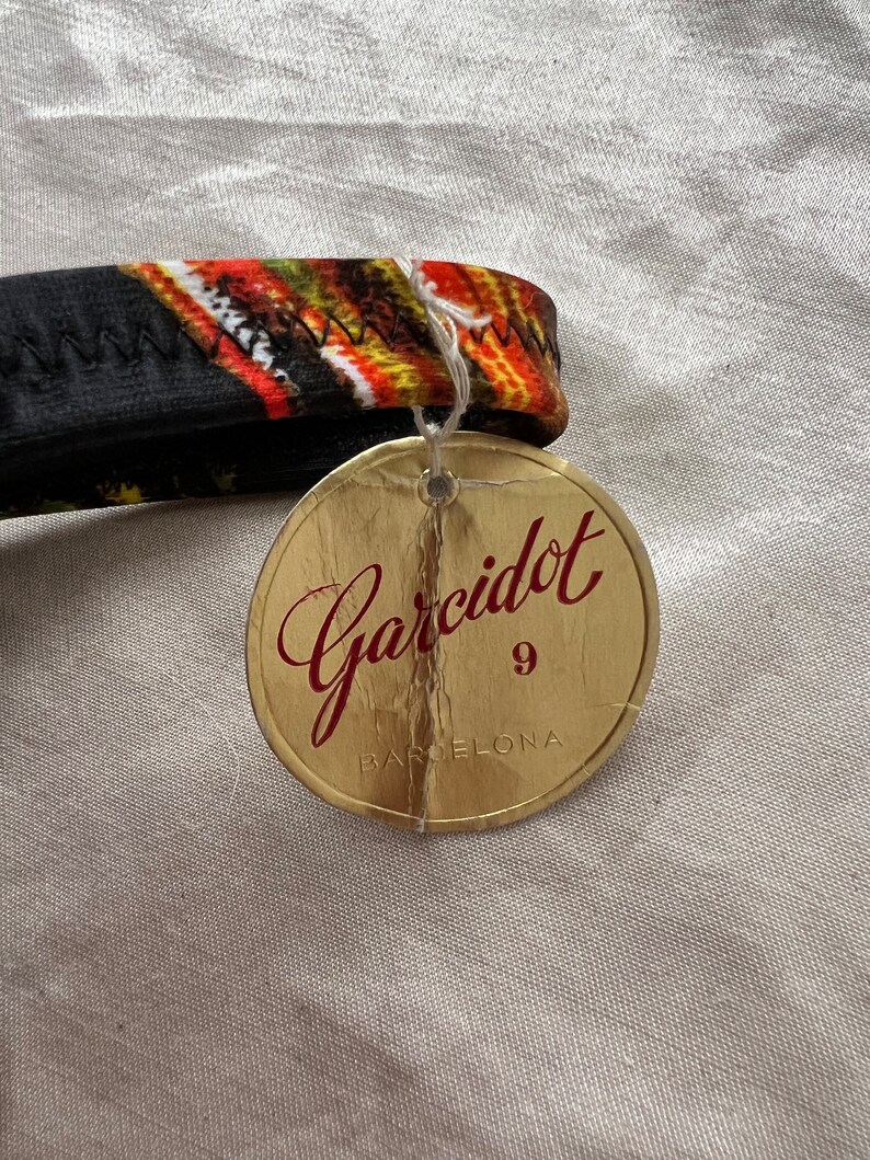 以下が含まれることがあります： 赤文字で「Garcidot 9 BARCELONA」と書かれた金色のタグ。タグはジグザグ模様の黒とオレンジの生地に付けられています。